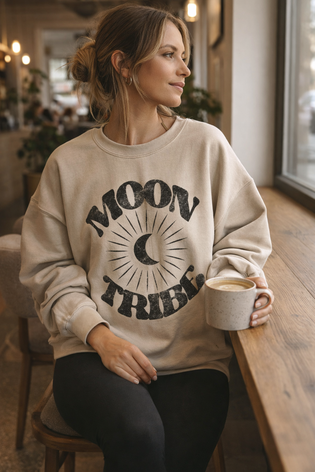 Woman wearing a 'Moon Tribe' sweatshirt holding a coffee mug in a casual setting.
