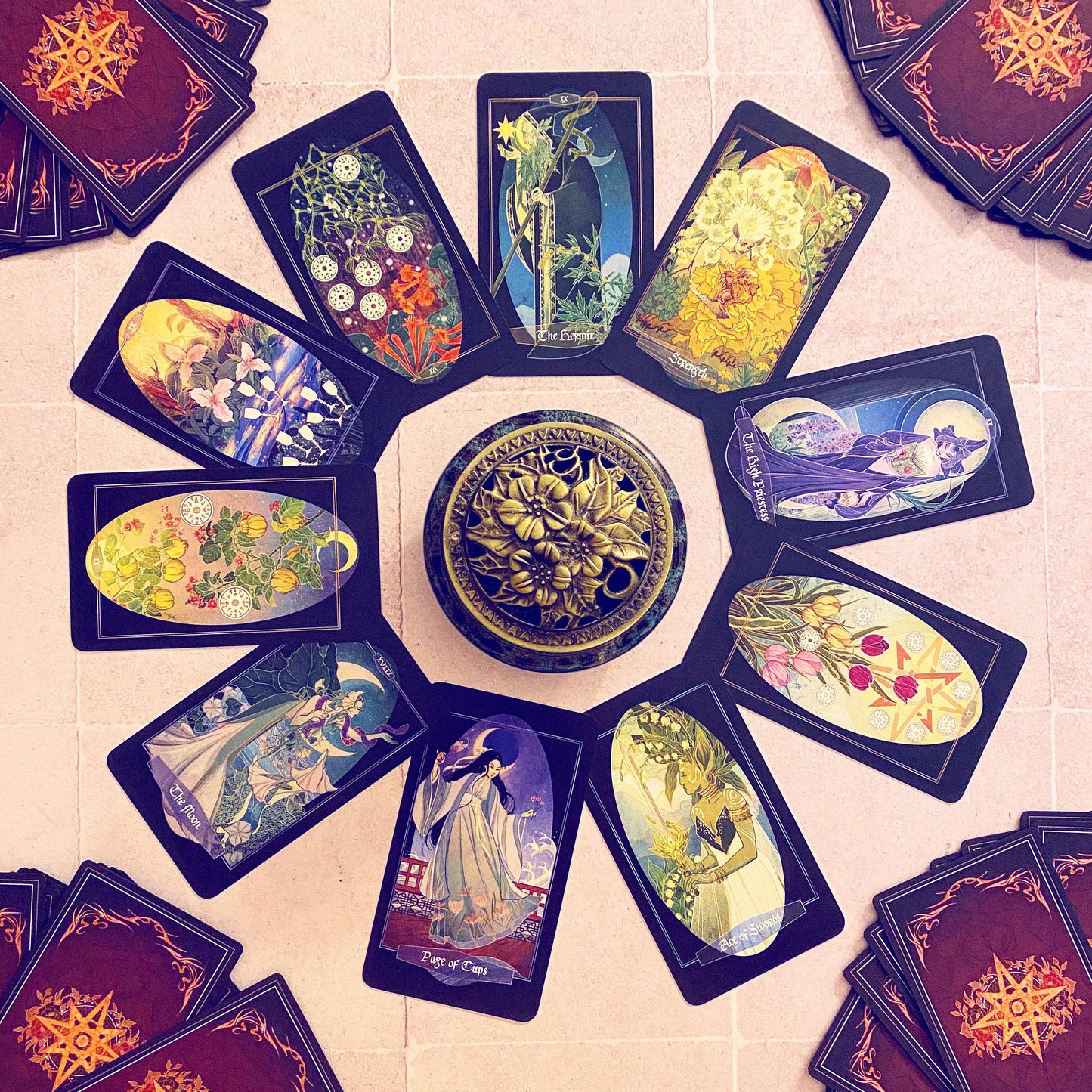 A circular arrangement of illustrated tarot or oracle cards with a decorative round box in the center, photographed from above on a light stone surface. The cards feature fantasy and floral artwork, and several card stacks are visible around the edges.