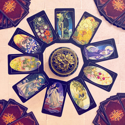 A circular arrangement of illustrated tarot or oracle cards with a decorative round box in the center, photographed from above on a light stone surface. The cards feature fantasy and floral artwork, and several card stacks are visible around the edges.