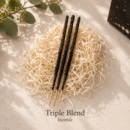 Three dark incense sticks resting on a bed of shredded natural paper, with the text 'Triple Blend Incense' below on a light textured surface with soft shadows and green leaves in the corner.