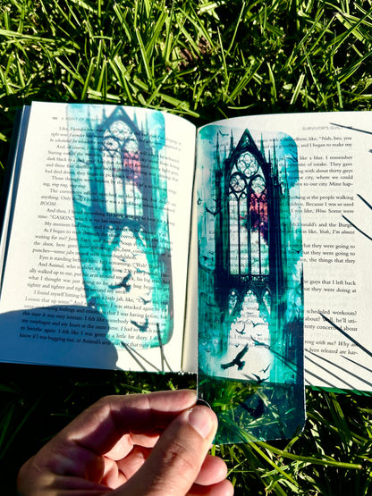 An open book held in a hand, showing printed text and a large artistic illustration of a gothic stained-glass window in teal, black, and red, set against a background of green grass in sunlight.