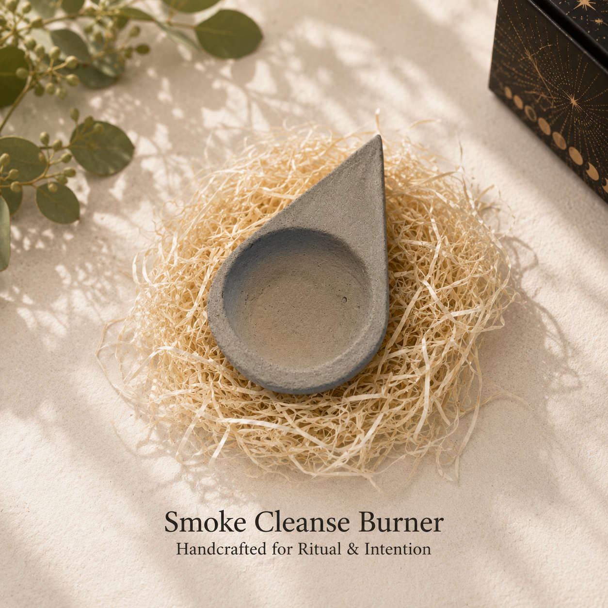 A teardrop-shaped gray ceramic smoke cleanse burner resting on a bed of natural jute fiber, with decorative green leaves and a black patterned box in the background. Text at the bottom reads 'Smoke Cleanse Burner' and 'Handcrafted for Ritual & Intention'.