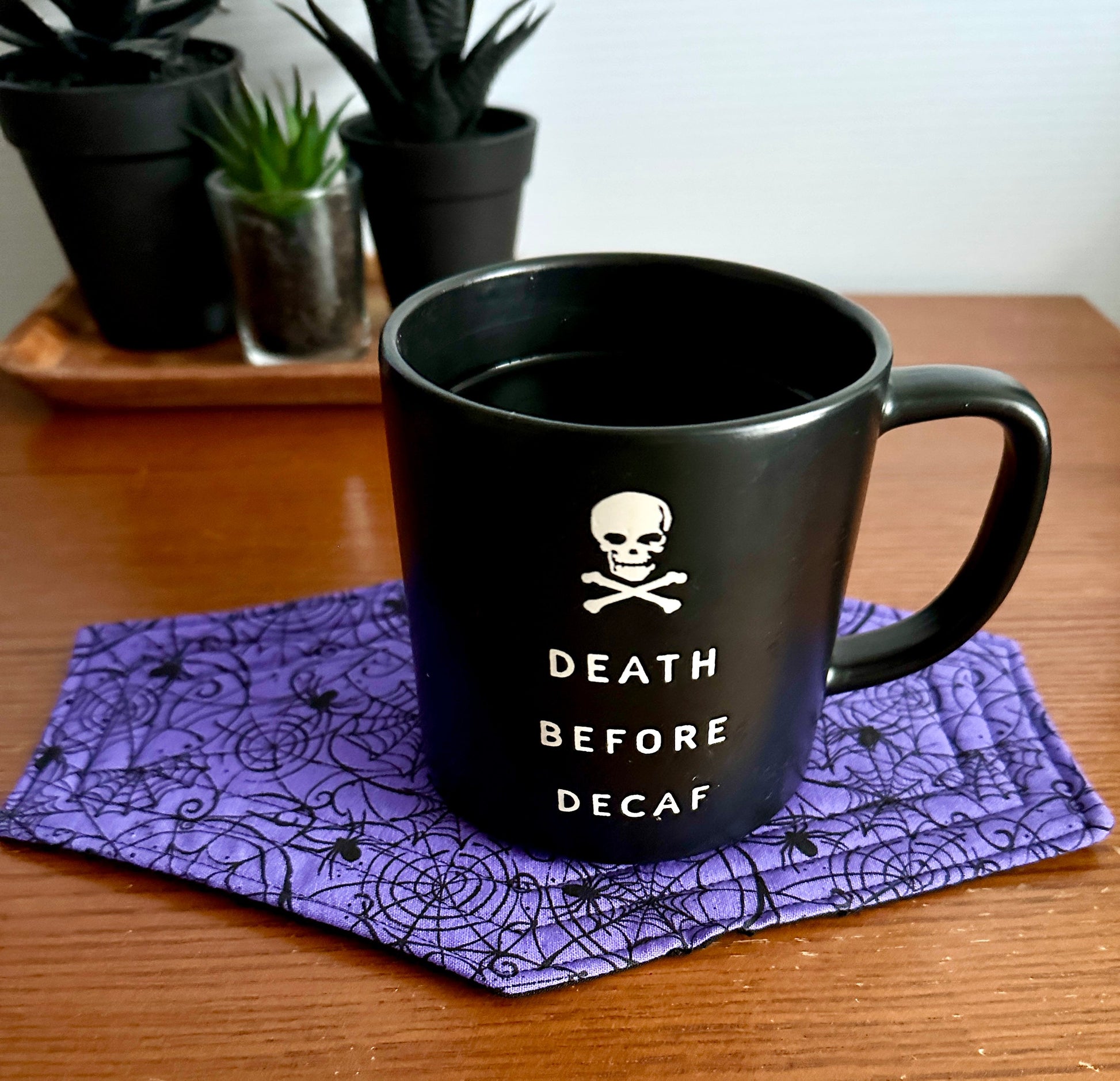 Black mug with 'Death Before Decaf' text on a purple spiderweb coaster, with plants in the background.
