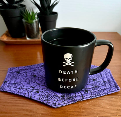 Black mug with 'Death Before Decaf' text on a purple spiderweb coaster, with plants in the background.