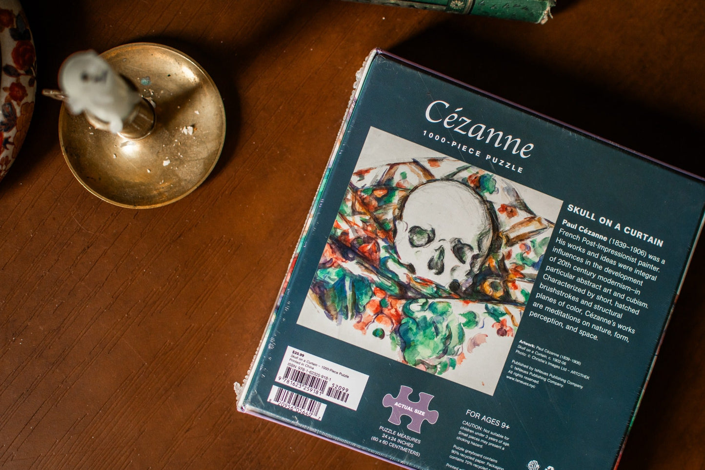 A boxed jigsaw puzzle titled “Cézanne: Skull on a Curtain” lies on a wooden tabletop. The package is dark teal with a large artwork panel showing a white skull over a colorful painted curtain scene.