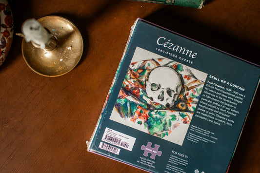 A boxed jigsaw puzzle titled “Cézanne: Skull on a Curtain” lies on a wooden tabletop. The package is dark teal with a large artwork panel showing a white skull over a colorful painted curtain scene.