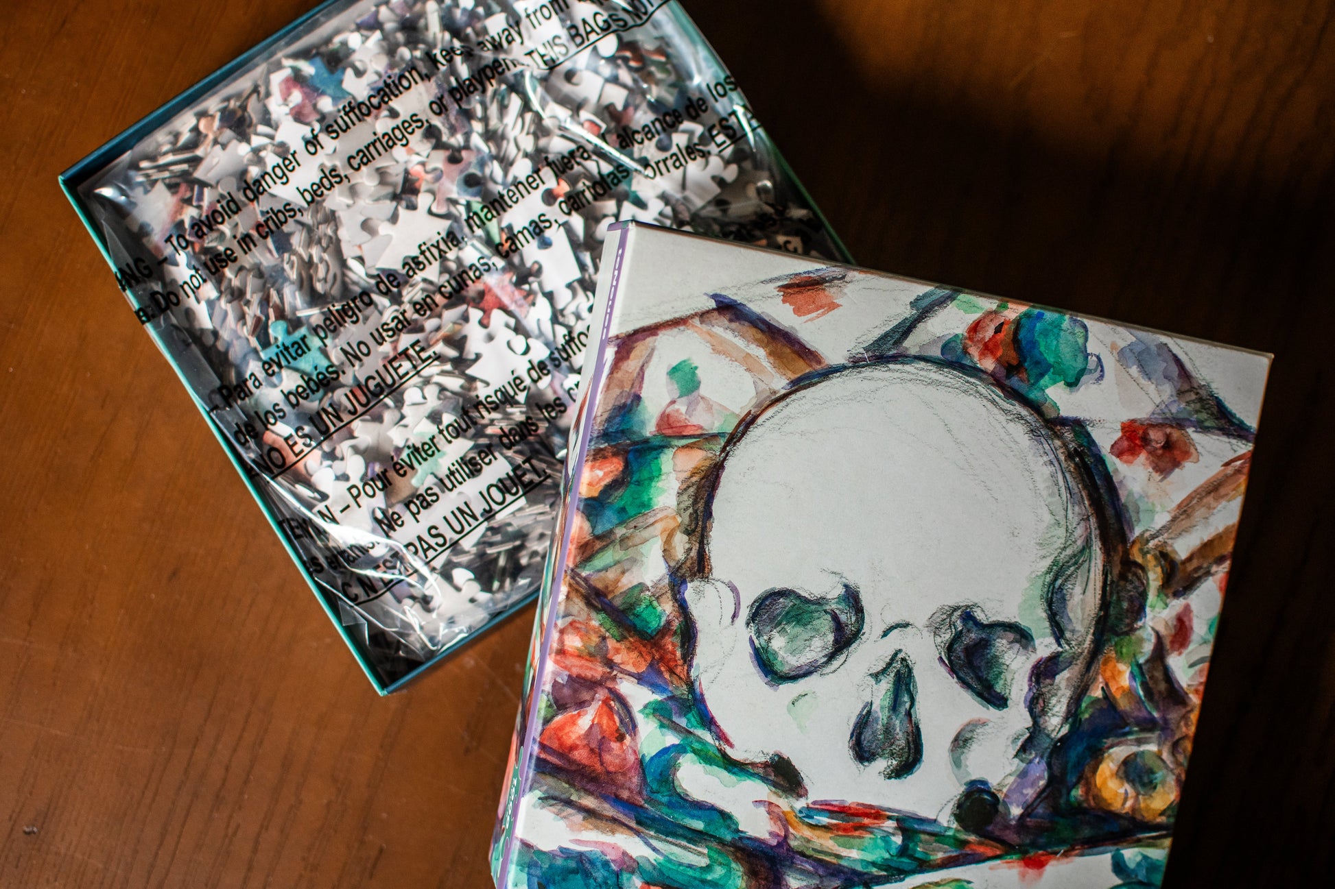 A square art print or card featuring a white skull illustration surrounded by colorful abstract brushstrokes, shown next to its open teal-edged packaging on a wooden surface.