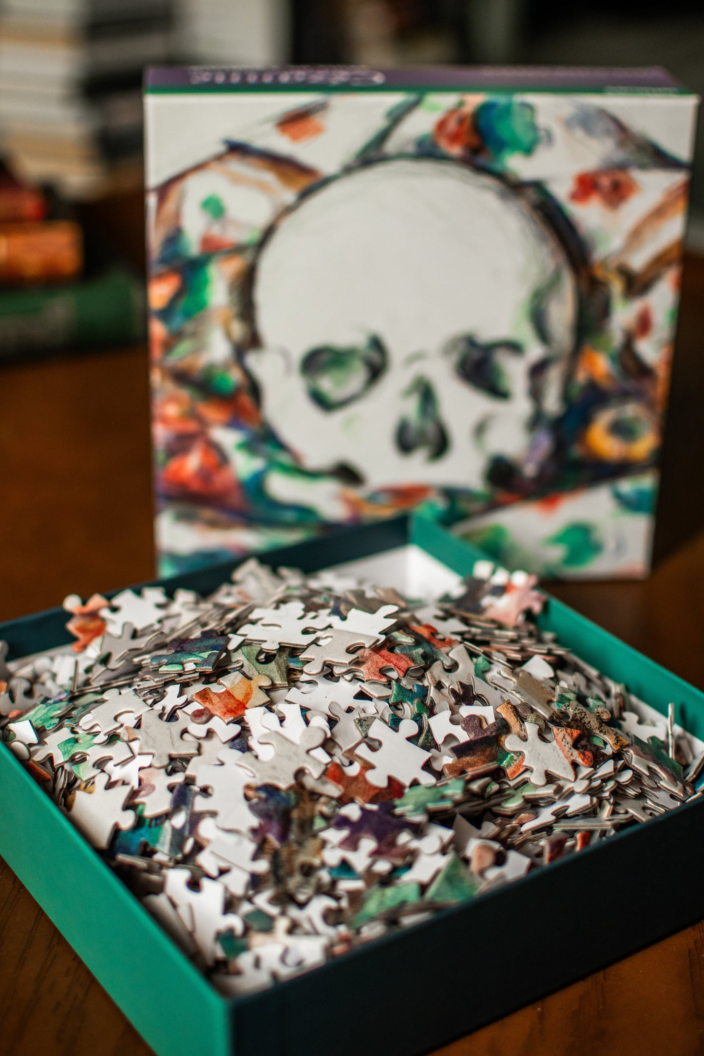 An open teal box filled with multicolored jigsaw puzzle pieces, with the illustrated puzzle box standing behind it on a wooden surface.