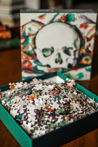 An open teal box filled with multicolored jigsaw puzzle pieces, with the illustrated puzzle box standing behind it on a wooden surface.