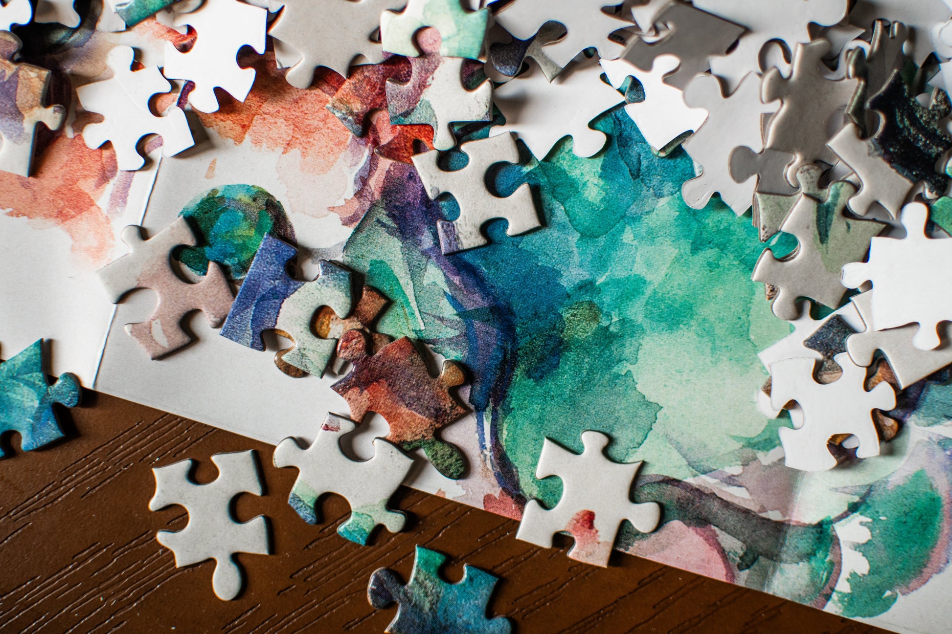 A close-up of a jigsaw puzzle with watercolor-style abstract artwork in teal, blue, purple, pink, and orange, with many loose white puzzle pieces scattered around it on a dark wooden surface.