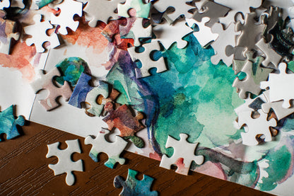 A close-up of a jigsaw puzzle with watercolor-style abstract artwork in teal, blue, purple, pink, and orange, with many loose white puzzle pieces scattered around it on a dark wooden surface.