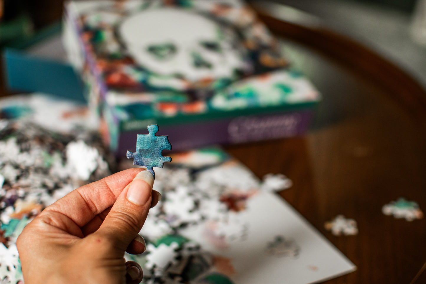 A hand holds a single light blue puzzle piece in front of a blurred jigsaw puzzle box and scattered puzzle pieces on a wooden table.