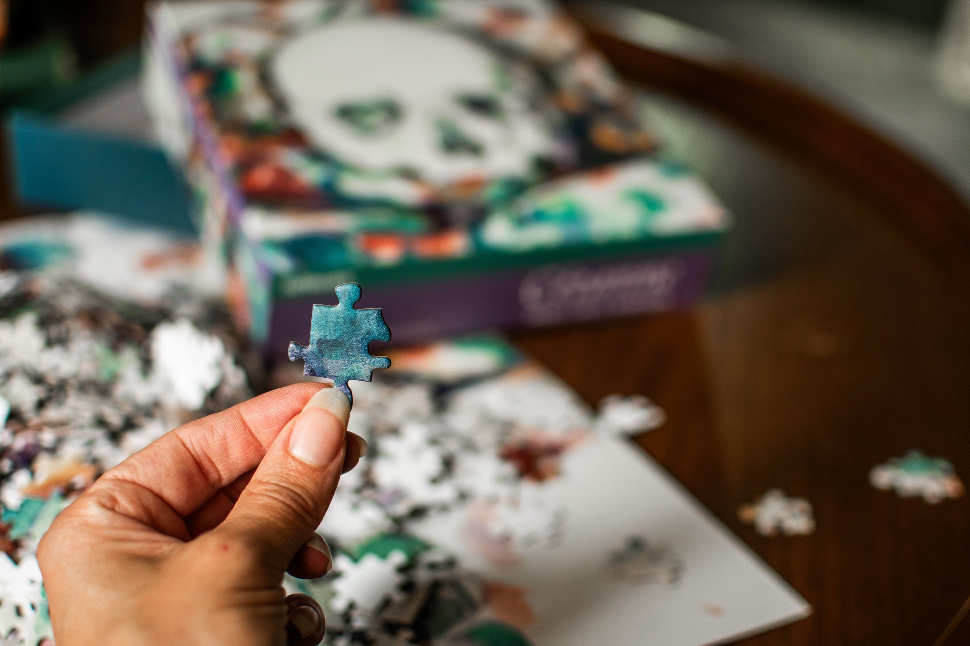 A hand holds a single light blue puzzle piece in front of a blurred jigsaw puzzle box and scattered puzzle pieces on a wooden table.
