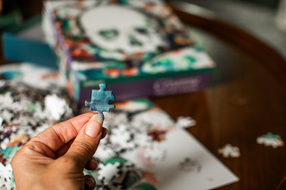 A hand holds a single light blue puzzle piece in front of a blurred jigsaw puzzle box and scattered puzzle pieces on a wooden table.