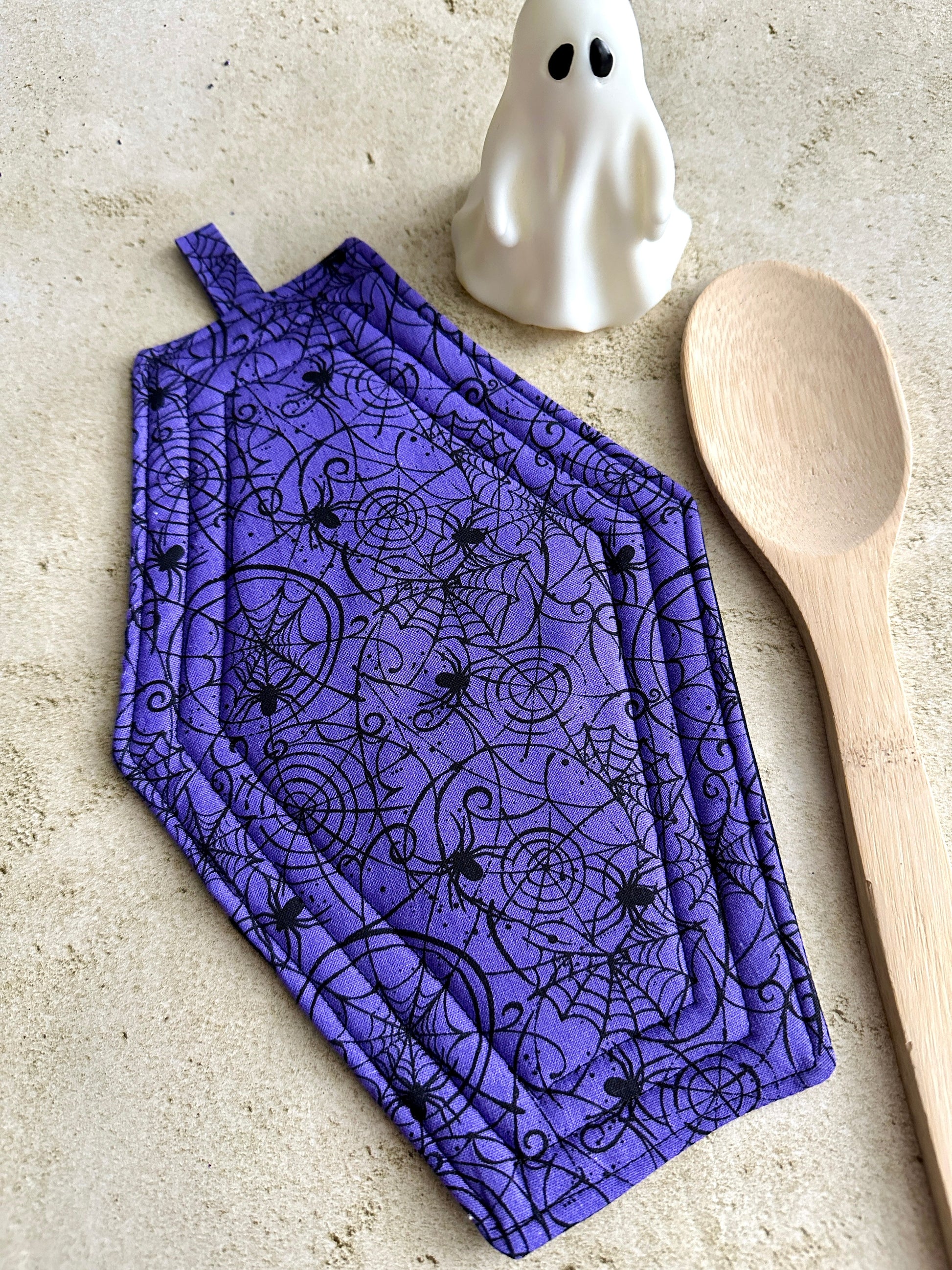 Purple oven mitt with Halloween design, wooden spoon, and ghost on a beige surface