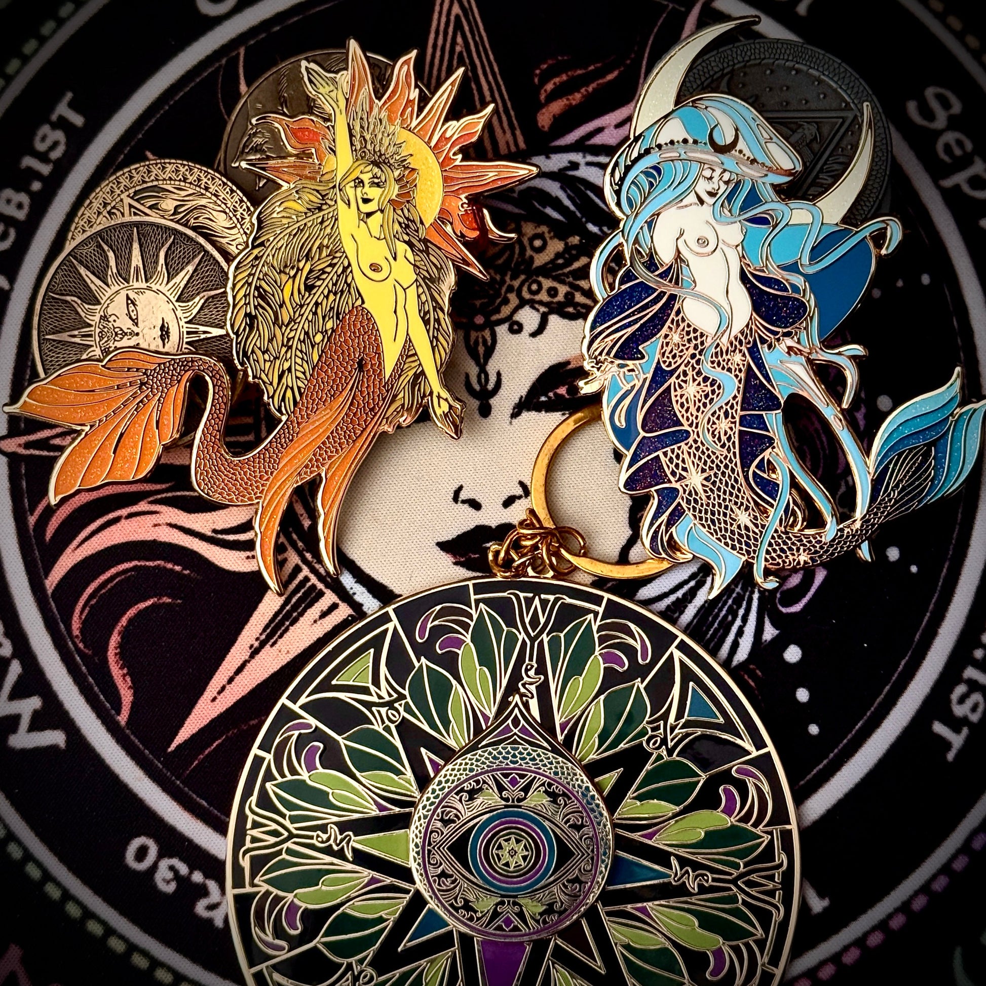 A set of mermaid-themed enamel pins arranged on a circular black background with celestial and mermaid artwork. The pins include a yellow sun-and-mermaid design, a blue-and-white mermaid with a crescent moon, and a large circular mermaid-and-sun motif pin at the bottom.