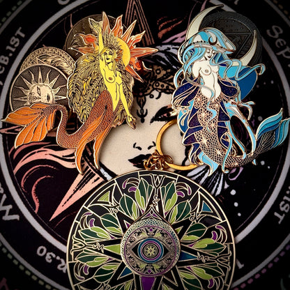 A set of mermaid-themed enamel pins arranged on a circular black background with celestial and mermaid artwork. The pins include a yellow sun-and-mermaid design, a blue-and-white mermaid with a crescent moon, and a large circular mermaid-and-sun motif pin at the bottom.