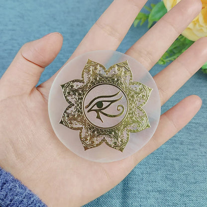 A hand holding a circular translucent silicone mold or coaster featuring a gold-toned lotus design with an eye of Horus symbol in the center, set against a blue textured background with blurred yellow flowers.