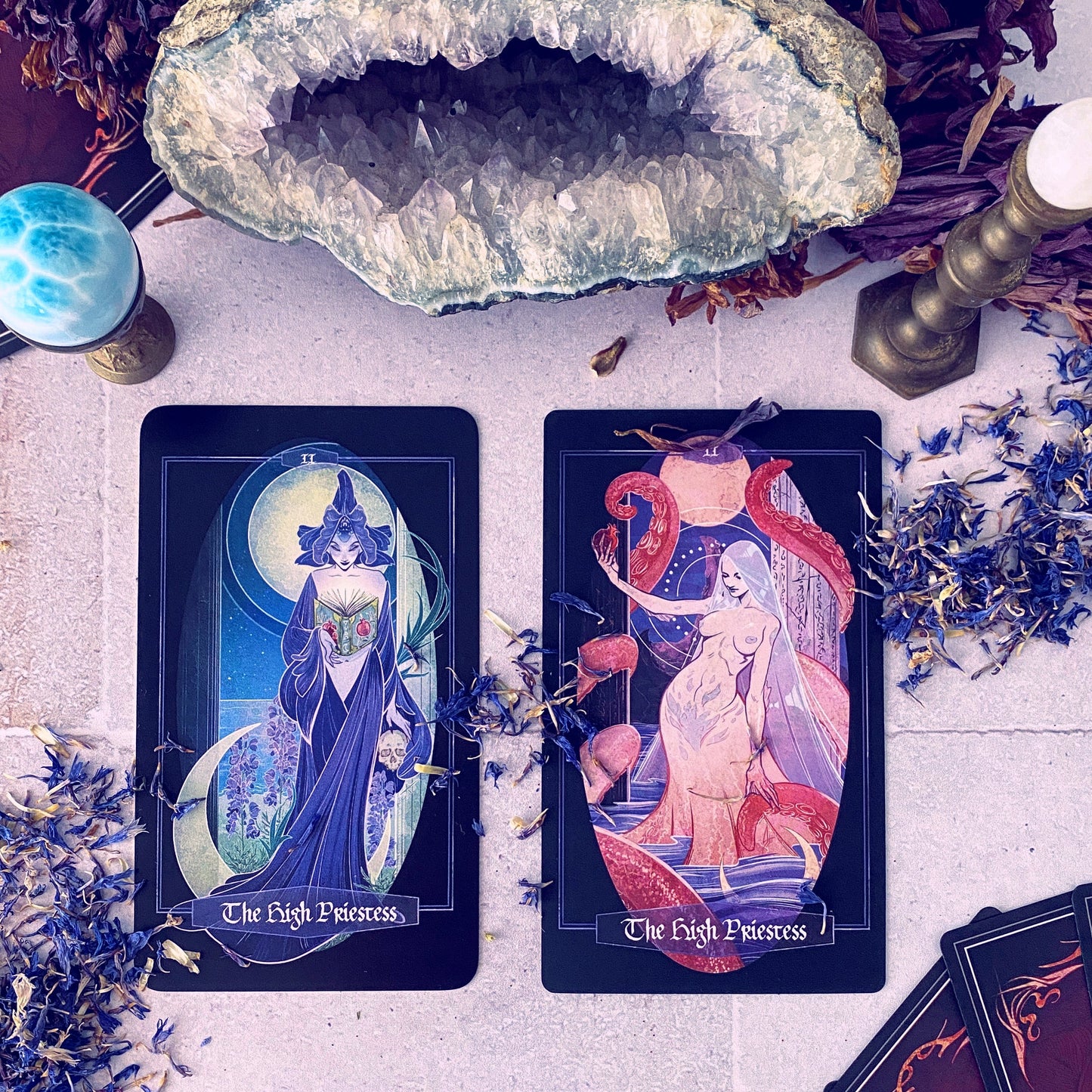 Two illustrated tarot cards labeled 'The High Priestess' are arranged on a light surface with dried flowers, crystals, a candle, and other mystical props around them.