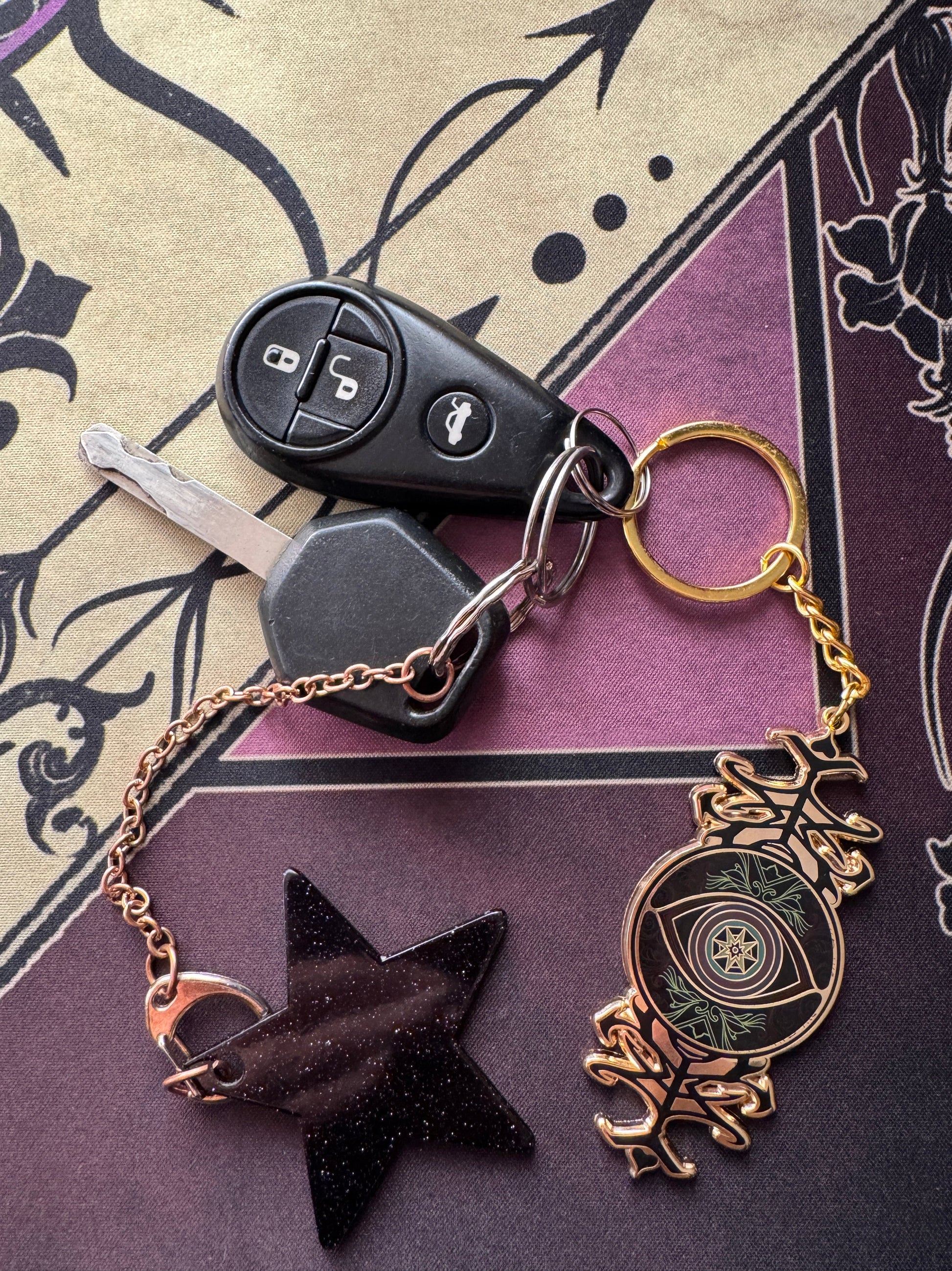 A keychain set displayed on a patterned surface, featuring a black car key fob, a gold-tone key ring, a black star-shaped charm, and a gold-tone circular charm with an eye of providence design.