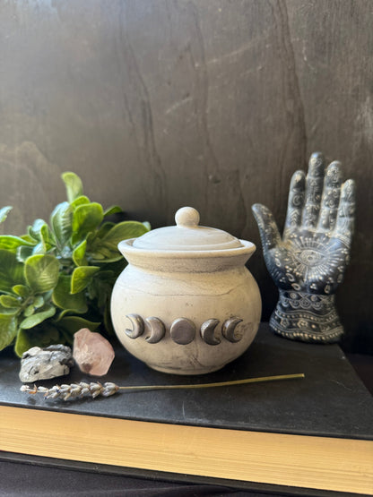 Decorative stone jar with moon phase symbols on a dark surface with plants and a palm reading hand.