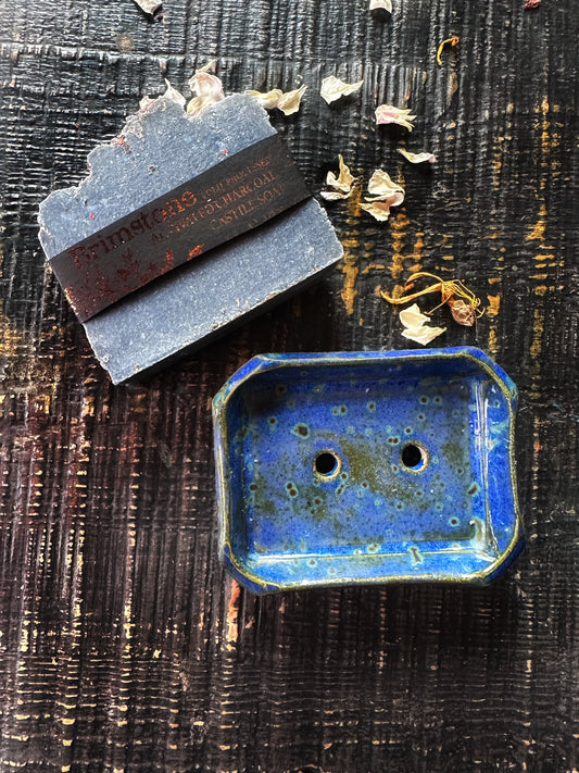 Bar of soap with a brand label on a textured dark surface next to a blue ceramic soap dish.