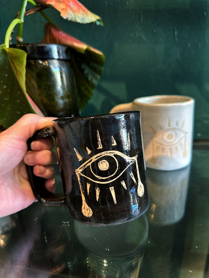 Black mug with gold eye design held by a hand, next to a white mug with a face design on a reflective surface.