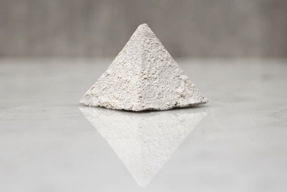 Pyramid-shaped object made of light gray material on a reflective surface with a neutral background