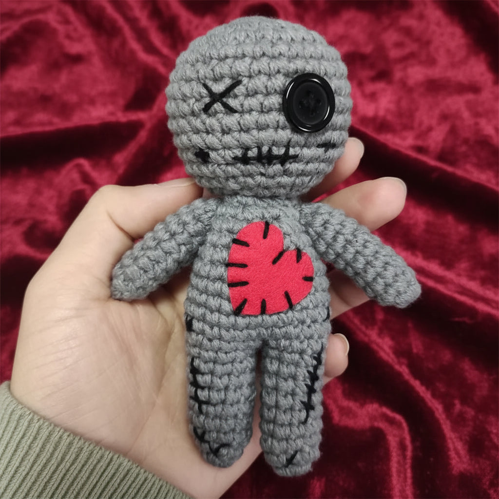 Gray crochet doll with a red heart held in a hand against a neutral background