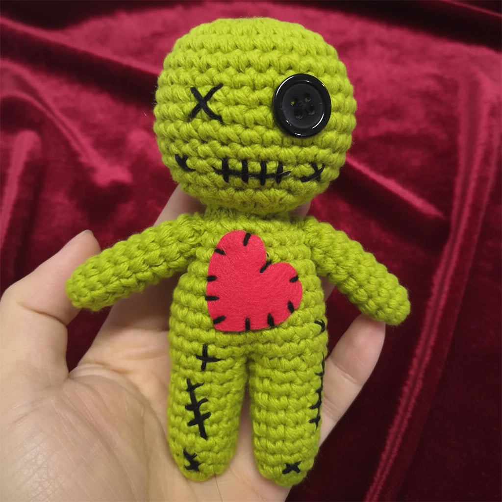 A small handmade crocheted doll held in a hand. The doll is lime green with black stitched details, a black button eye, and a red felt heart on the chest.
