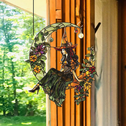 Decorative stained glass panel with a witch sitting on a moon, surrounded by flowers, hanging in front of a window with a view of trees.