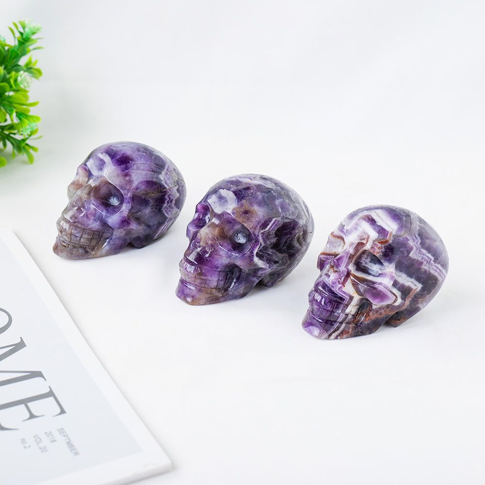 Three purple crystal skulls on a white surface with a plant in the background
