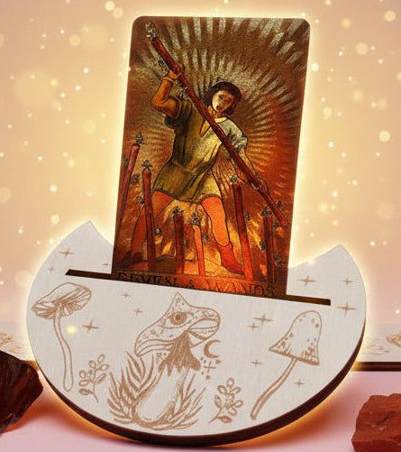Card with an angel holding a cross on a decorative stand with mushroom illustrations, candles in the background.