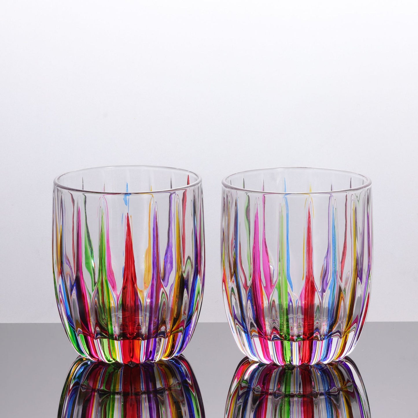 Two colorful glass tumblers with multicolored stripes on a reflective surface.