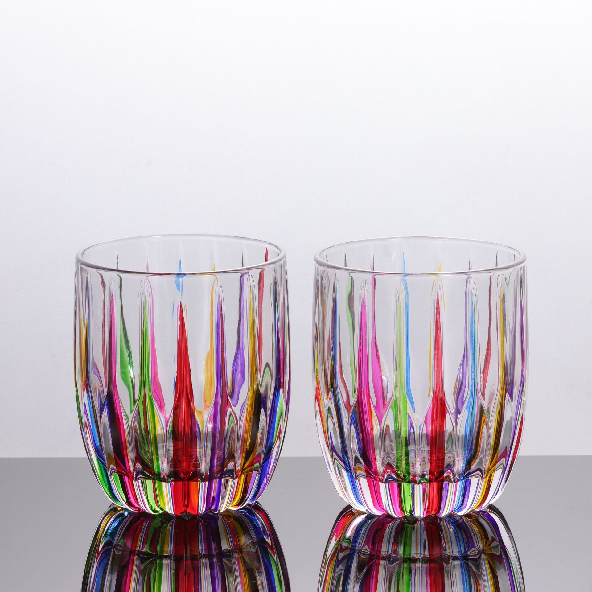 Two colorful glass tumblers with multicolored stripes on a reflective surface.