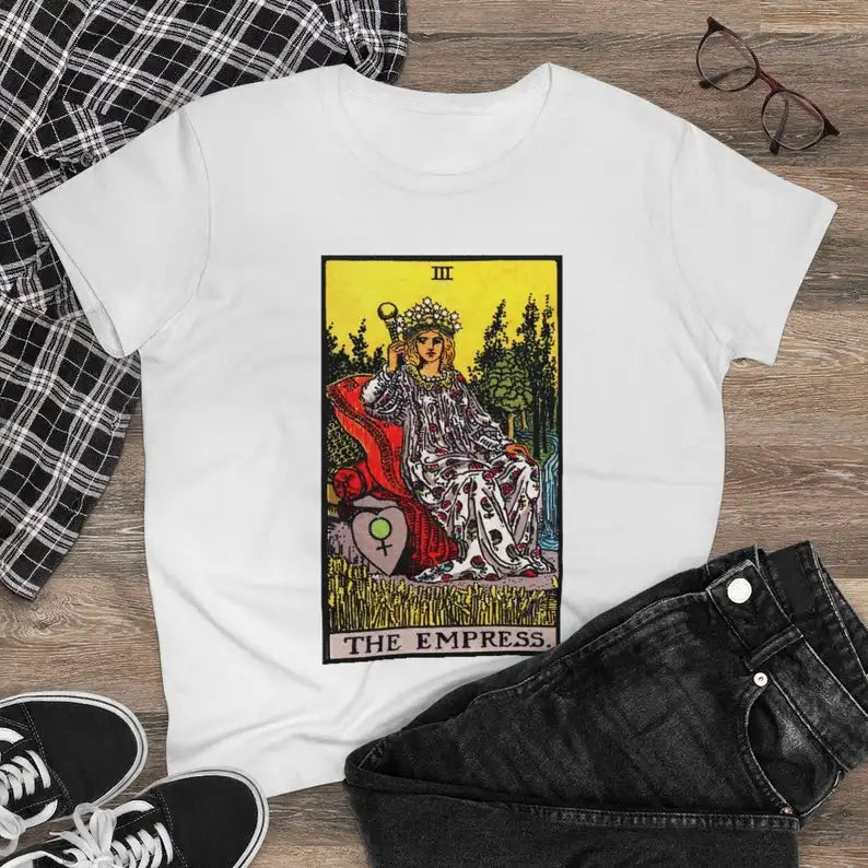 White t-shirt with 'The Empress' tarot card design on a wooden surface with plaid fabric, black pants, and sneakers.