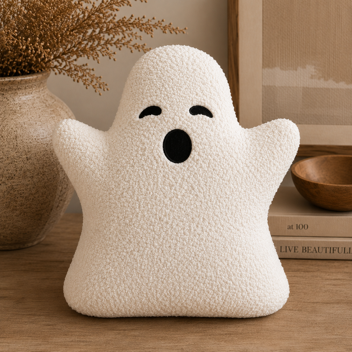 A white textured plush ghost pillow with embroidered black eyes and mouth, sitting on a wooden table in a cozy room setting with a vase, books, and a bowl in the background.