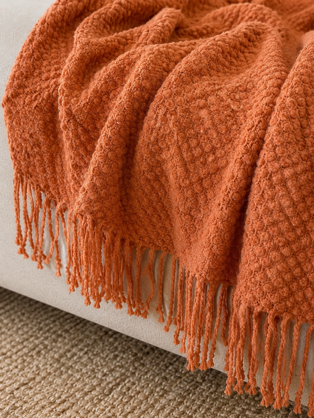 A close-up of a textured terracotta orange throw blanket with fringe, draped over a light beige upholstered armchair on a woven rug.
