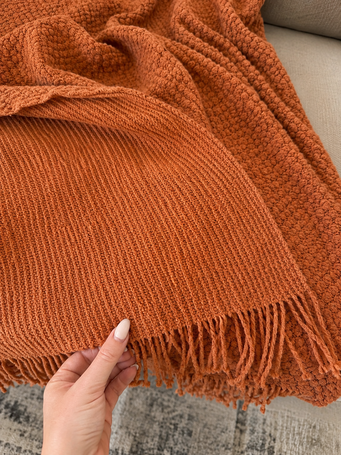 A close-up of a chunky knit throw blanket in a warm rust orange color, draped over a light-colored sofa with a hand holding the fringed edge.