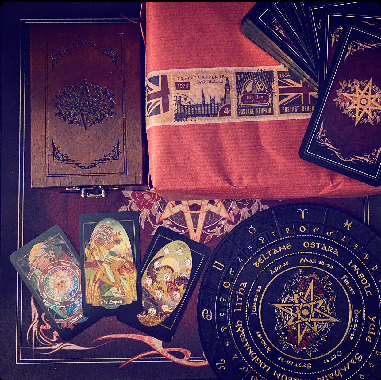 A styled flat-lay of tarot card and occult-themed items on a dark surface: a brown embossed book or journal, a folded orange-red cloth with vintage stamps, a stack of dark tarot cards, several illustrated cards, and a large circular black pentagram wheel with gold text and symbols.