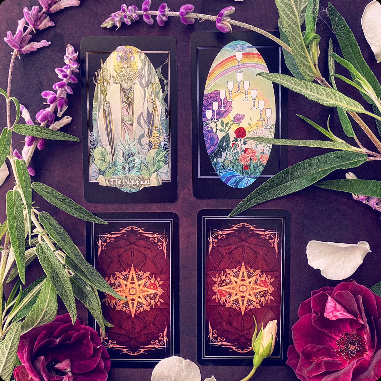 Four tarot cards arranged in a 2x2 grid on a dark purple background, surrounded by decorative botanical elements including purple flowers, green leaves, rose petals, and a white rose bud. Two cards are shown face up with illustrated fantasy and landscape scenes, and two are shown face down with ornate red and gold backs.