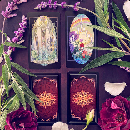 Four tarot cards arranged in a 2x2 grid on a dark purple background, surrounded by decorative botanical elements including purple flowers, green leaves, rose petals, and a white rose bud. Two cards are shown face up with illustrated fantasy and landscape scenes, and two are shown face down with ornate red and gold backs.