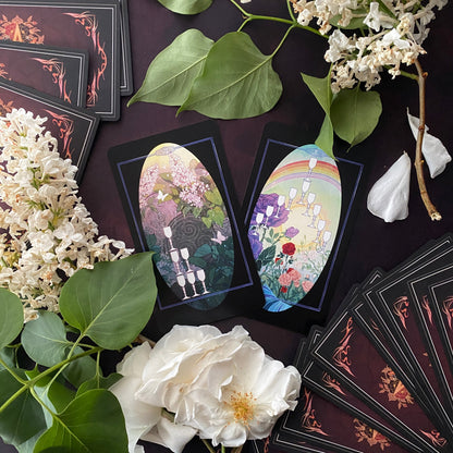 A styled flat-lay of a tarot card deck with two illustrated cards displayed upright and several cards fanned out, surrounded by white flowers and green leaves on a dark background.