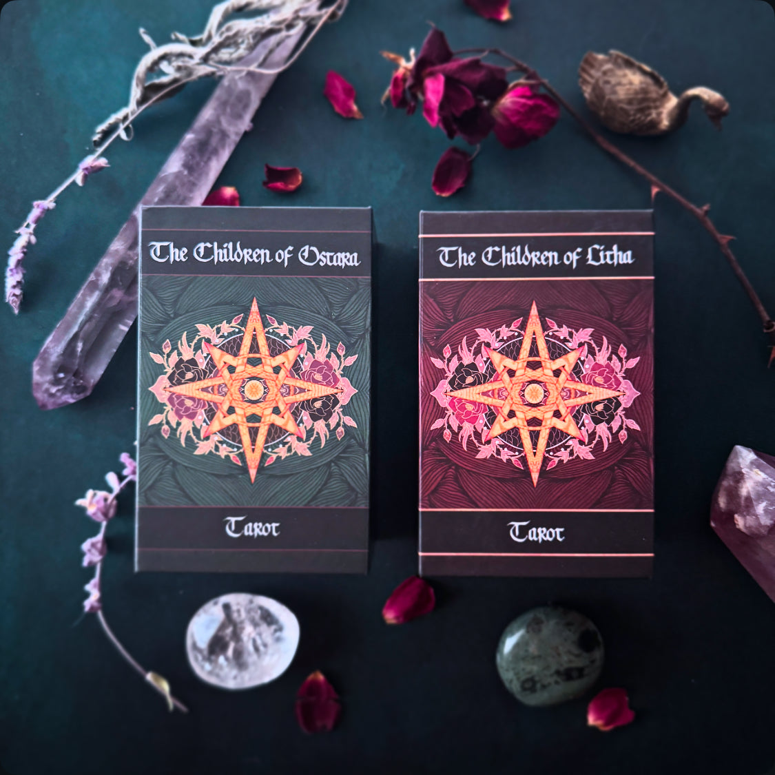 Two tarot card boxes from The Children of Ocuma and The Children of Lichu arranged on a dark surface with crystals, dried flowers, and rose petals around them.