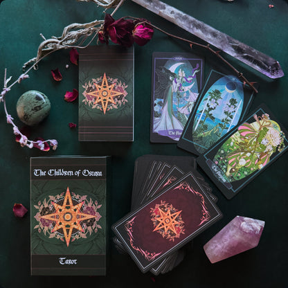 Wiccan tarot deck titled 'The Children of Ororo' arranged on a dark green surface with crystals, rose petals, and dried branches. The image shows the tarot box, a sealed deck, a fanned deck, and two cards with detailed fantasy illustrations.