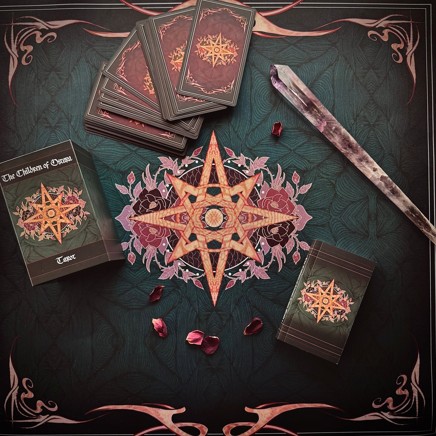 A styled flat-lay of a tarot card deck set on a dark decorative surface. Visible items include the tarot box labeled 'The Children of Osiris Tarot', several fanned tarot cards, a large central star-and-floral design, a crystal wand, and a smaller tarot card pack.