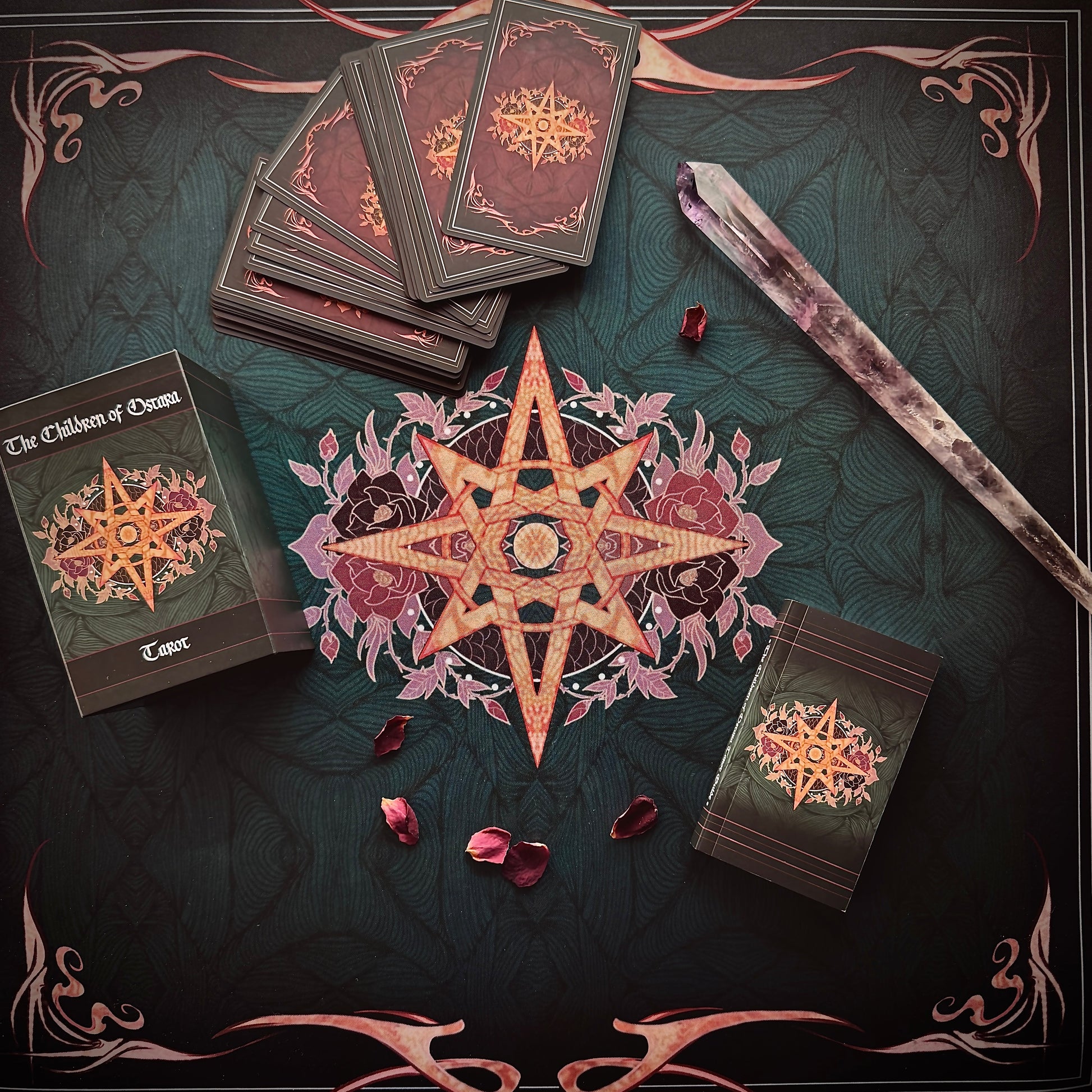 A styled flat-lay of a tarot card deck set on a dark decorative surface. Visible items include the tarot box labeled 'The Children of Osiris Tarot', several fanned tarot cards, a large central star-and-floral design, a crystal wand, and a smaller tarot card pack.