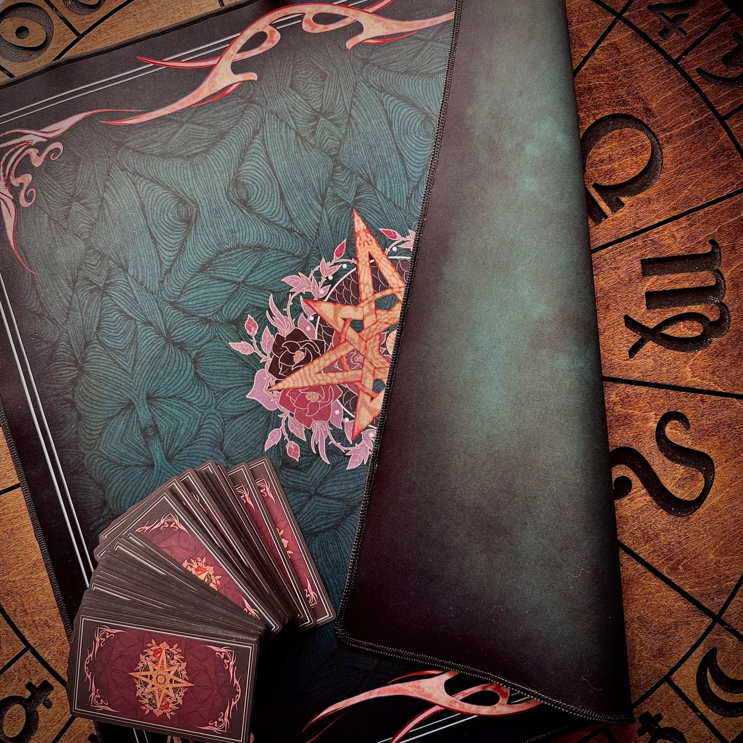 A dark teal and black leather spellcasting playmat with a central pentagram design, shown alongside a fanned deck of cards with red backs and a brown parchment-style board with astrological symbols.