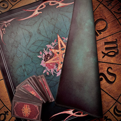 A dark teal and black leather spellcasting playmat with a central pentagram design, shown alongside a fanned deck of cards with red backs and a brown parchment-style board with astrological symbols.