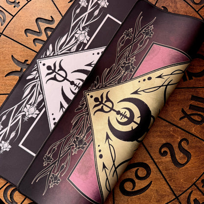 Two decorative rectangular mats or pads with occult-style graphics, shown partially rolled on a brown printed surface. One mat is black with white floral and geometric motifs, and the other is dark pink and beige with black symbols and floral line art.
